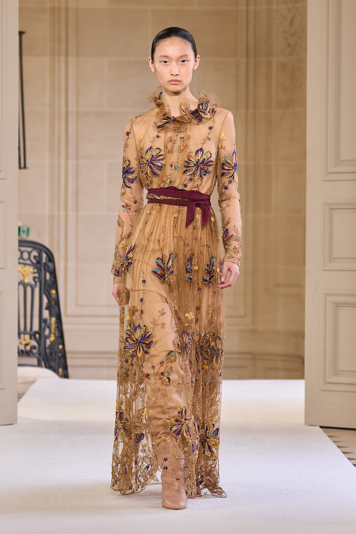 Model on a runway wearing a long sheer beige gown with blue and gold floral embroidery and a burgundy sash at the waist.
