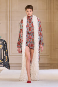 Model walking a runway wearing a red and black patterned romper with a long white faux‑fur scarf.