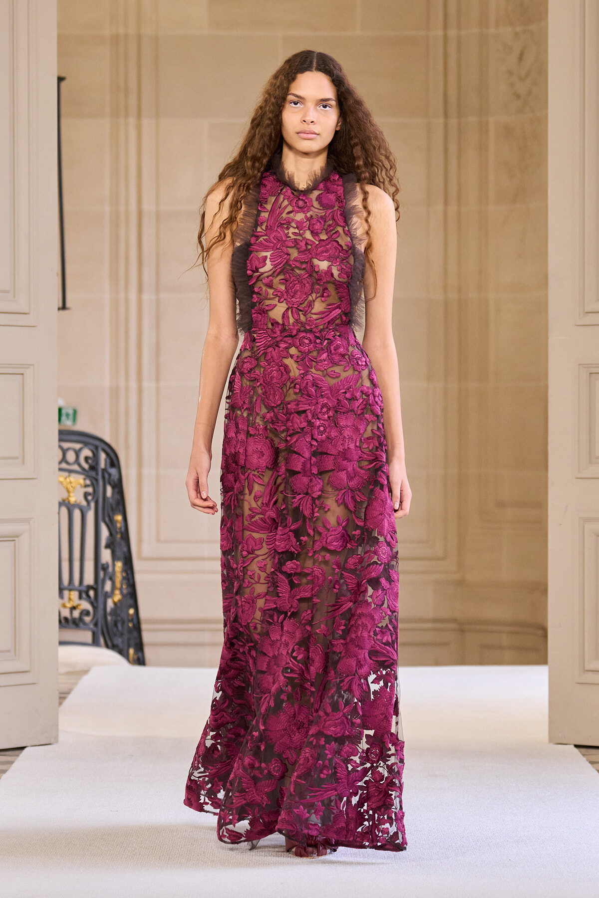 Model walking a runway in a floor-length magenta lace gown with floral embroidery and sheer panels in a beige, elegant venue.