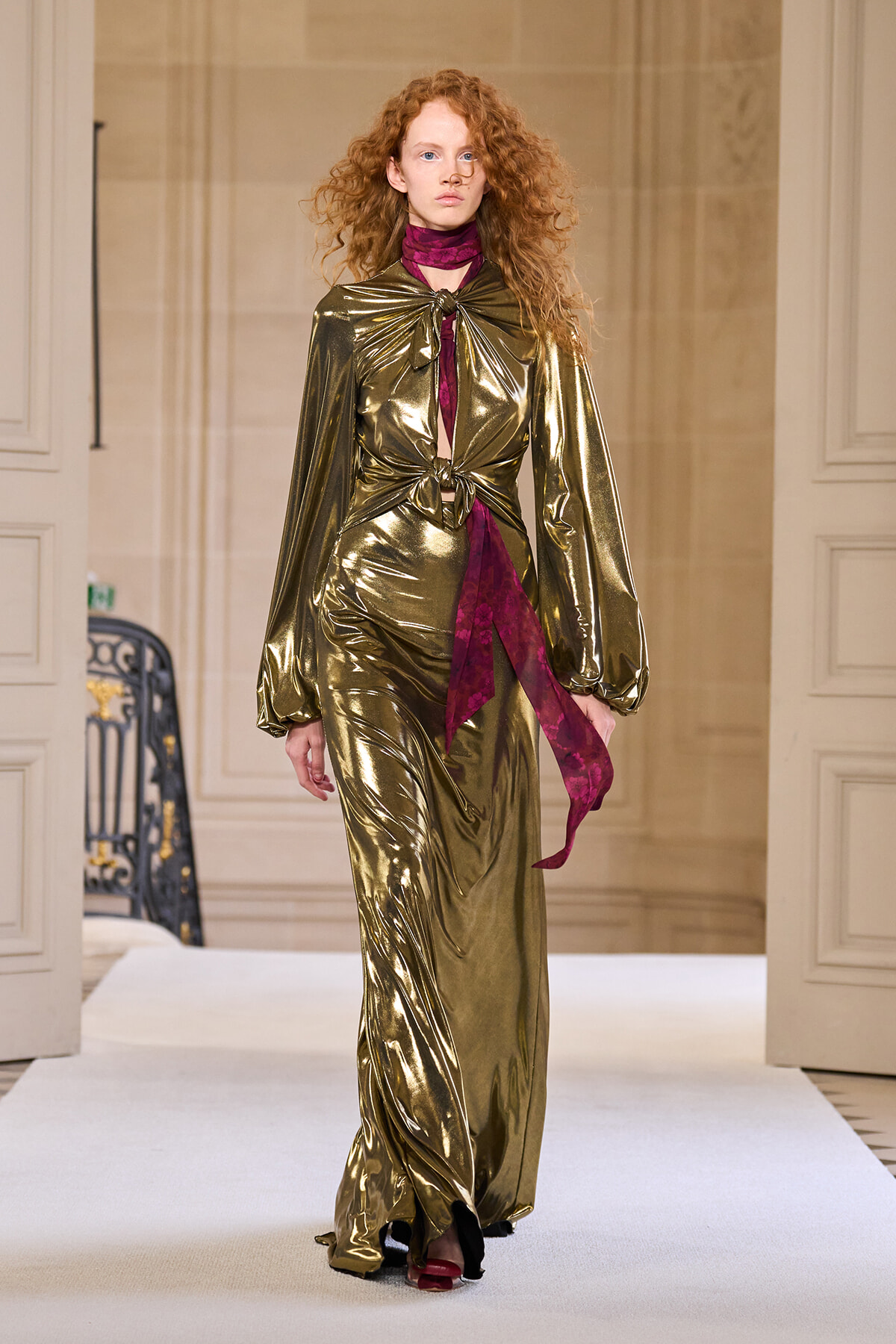 Model walks a runway in a metallic gold jumpsuit-like gown with a knotted front and magenta scarf.