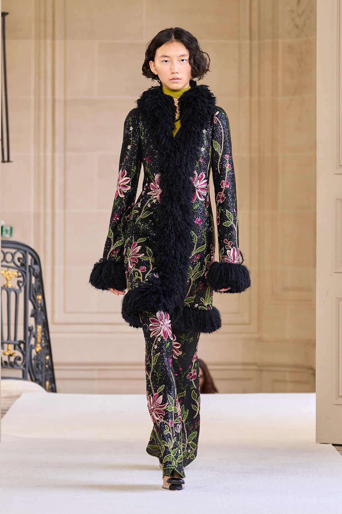 Model walks runway in a black velvet ensemble with bold pink and green floral embroidery and chunky black faux-fur trim at the collar and cuffs.