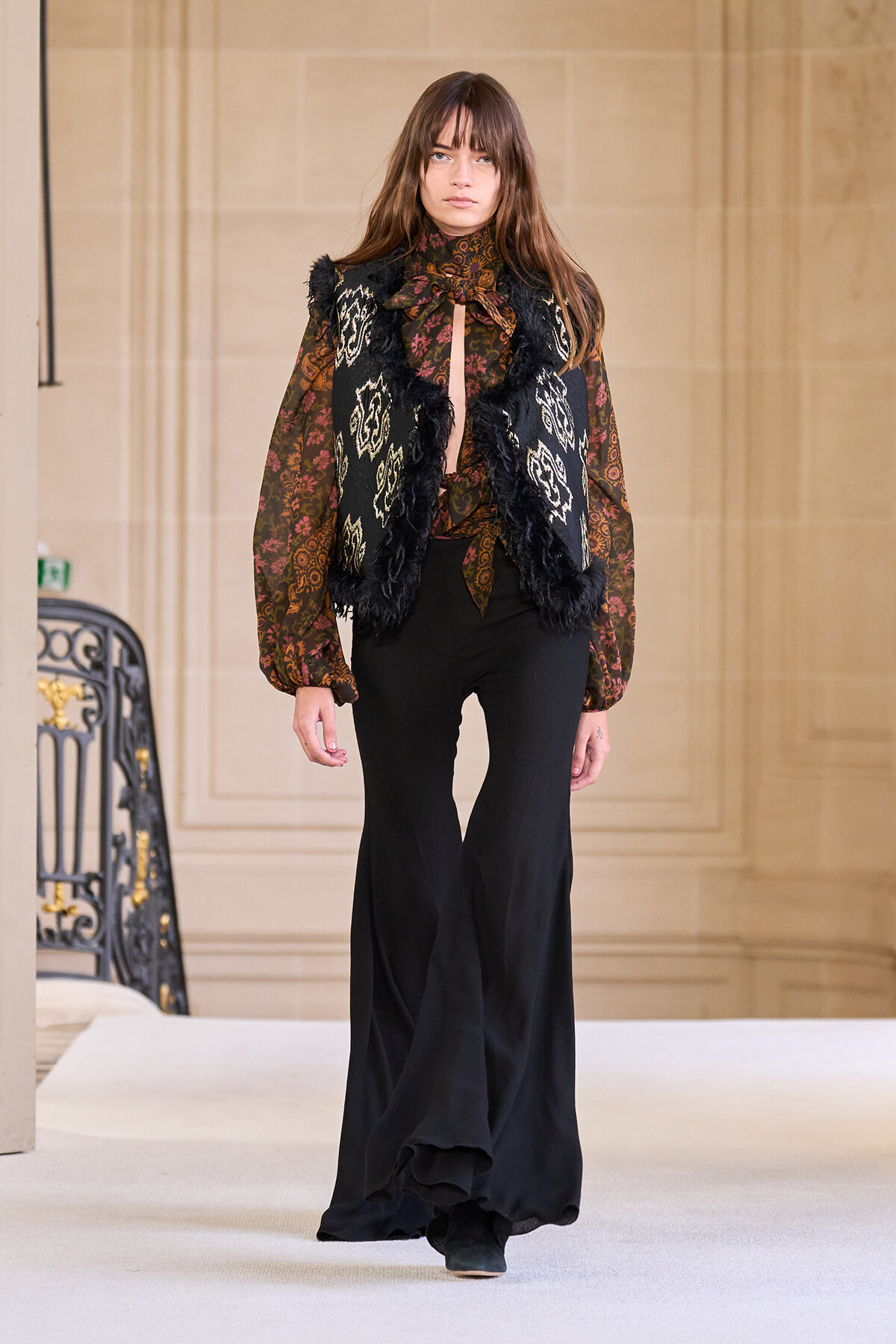 Model wearing a sheer burgundy patterned blouse with a bow, a black fringed fur-trim vest, and wide-leg black pants on a runway.