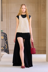 Model on a runway wearing a cream knit sleeveless top over a black draped skirt, carrying a red woven handbag.