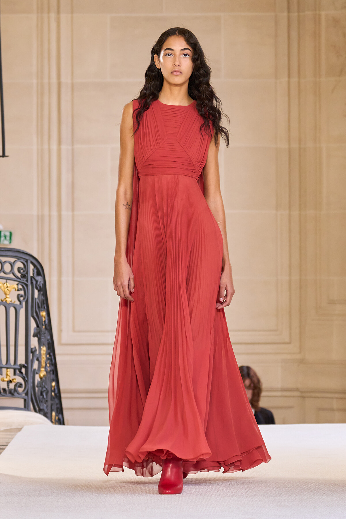 Model on a runway wearing a floor-length red chiffon gown with a structured, pleated bodice and flowing skirt, walking the catwalk.