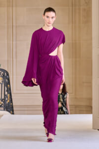 Model walking the runway in a magenta draped jumpsuit with an asymmetrical cape sleeve and waist cutout.
