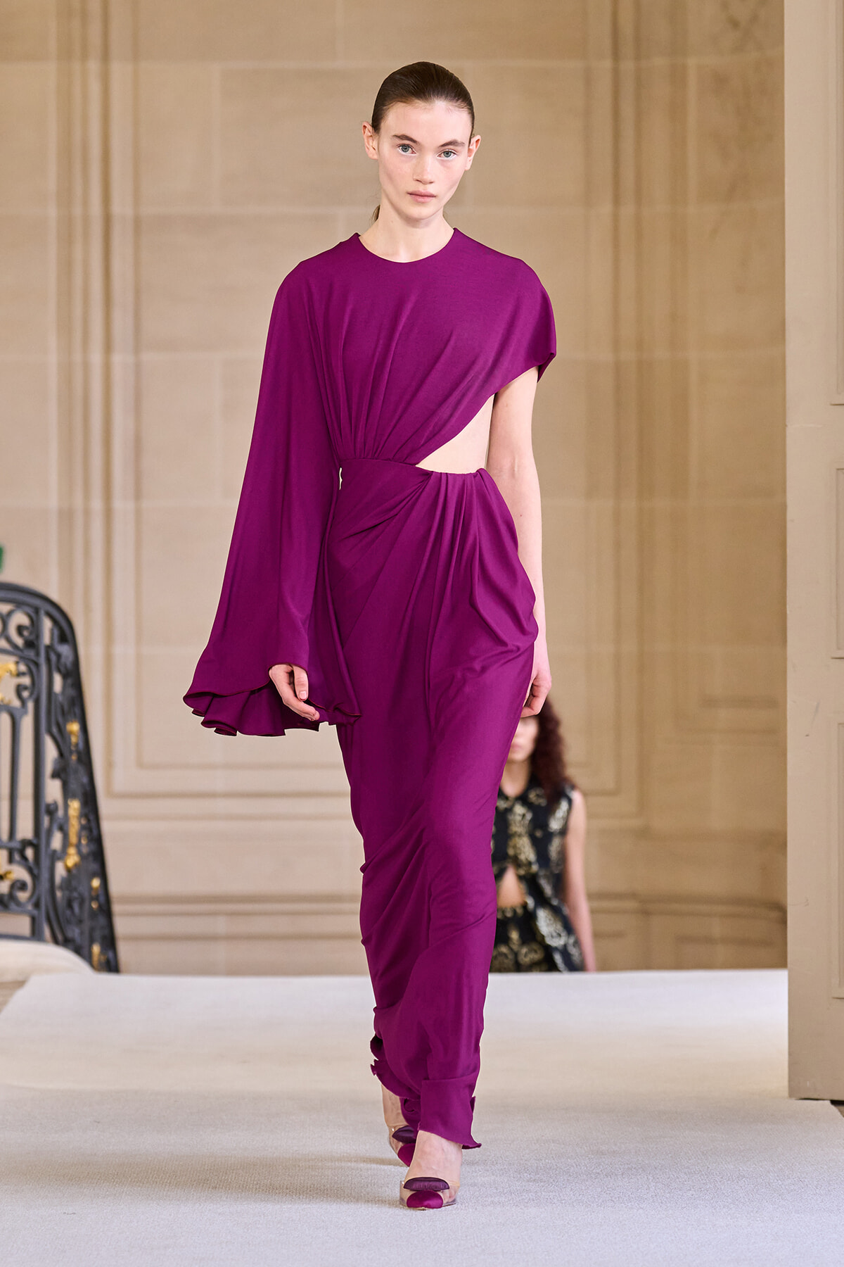 Model walking the runway in a magenta draped jumpsuit with an asymmetrical cape sleeve and waist cutout.