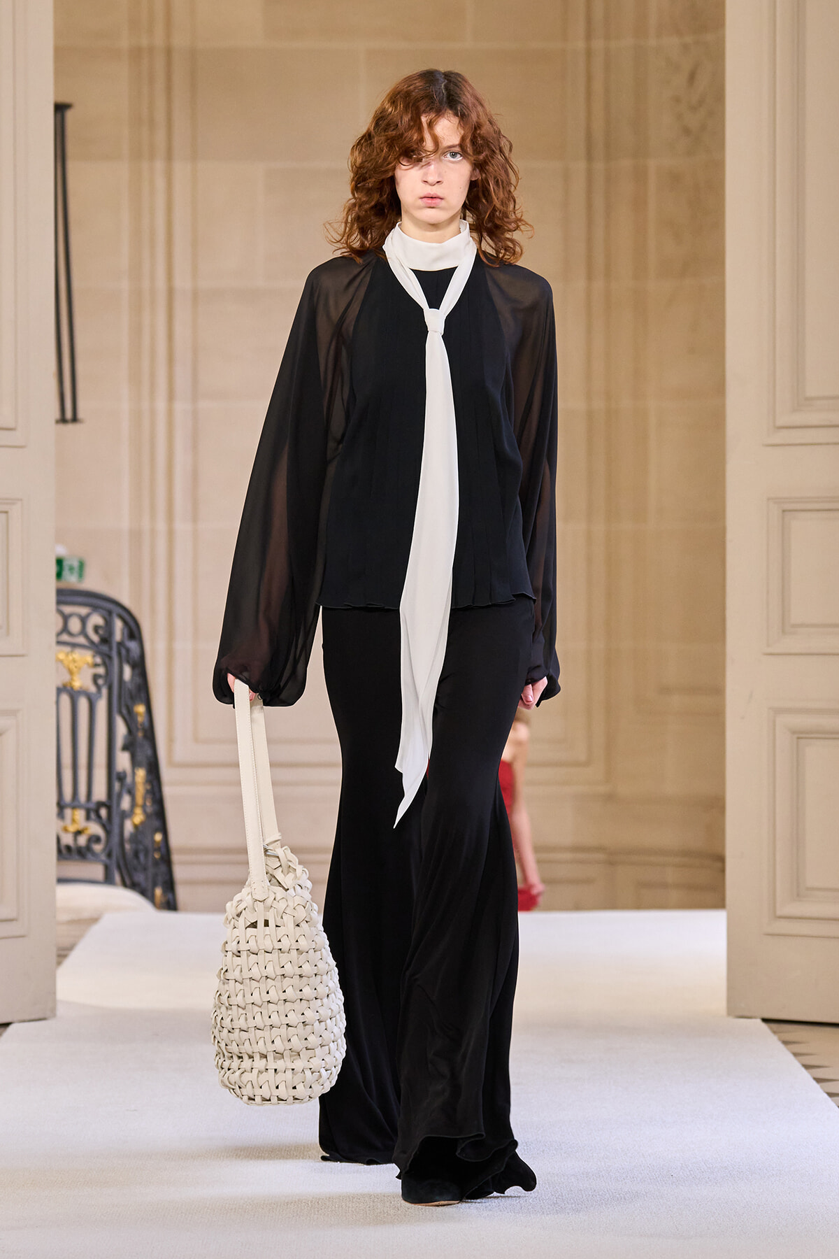 Model walking a runway in a sheer black blouse with a long white scarf and black floor-length skirt, carrying a white woven bag.