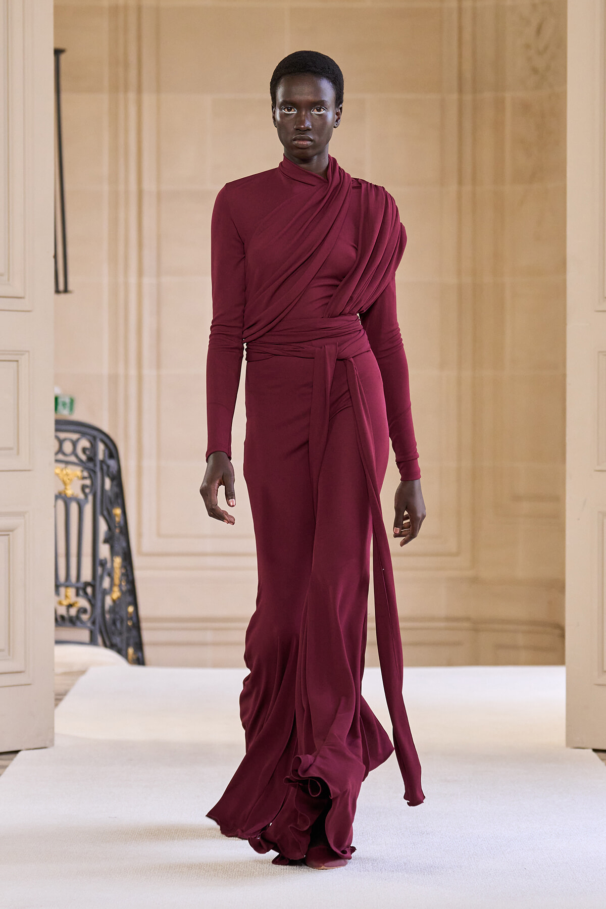 Model walks the runway in a burgundy draped wrap dress with long sleeves and a tied waist.