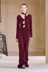 Runway model wearing a burgundy long-sleeve gown with cutouts at the midriff and a tied scarf-like neckpiece, on a neutral fashion show backdrop.
