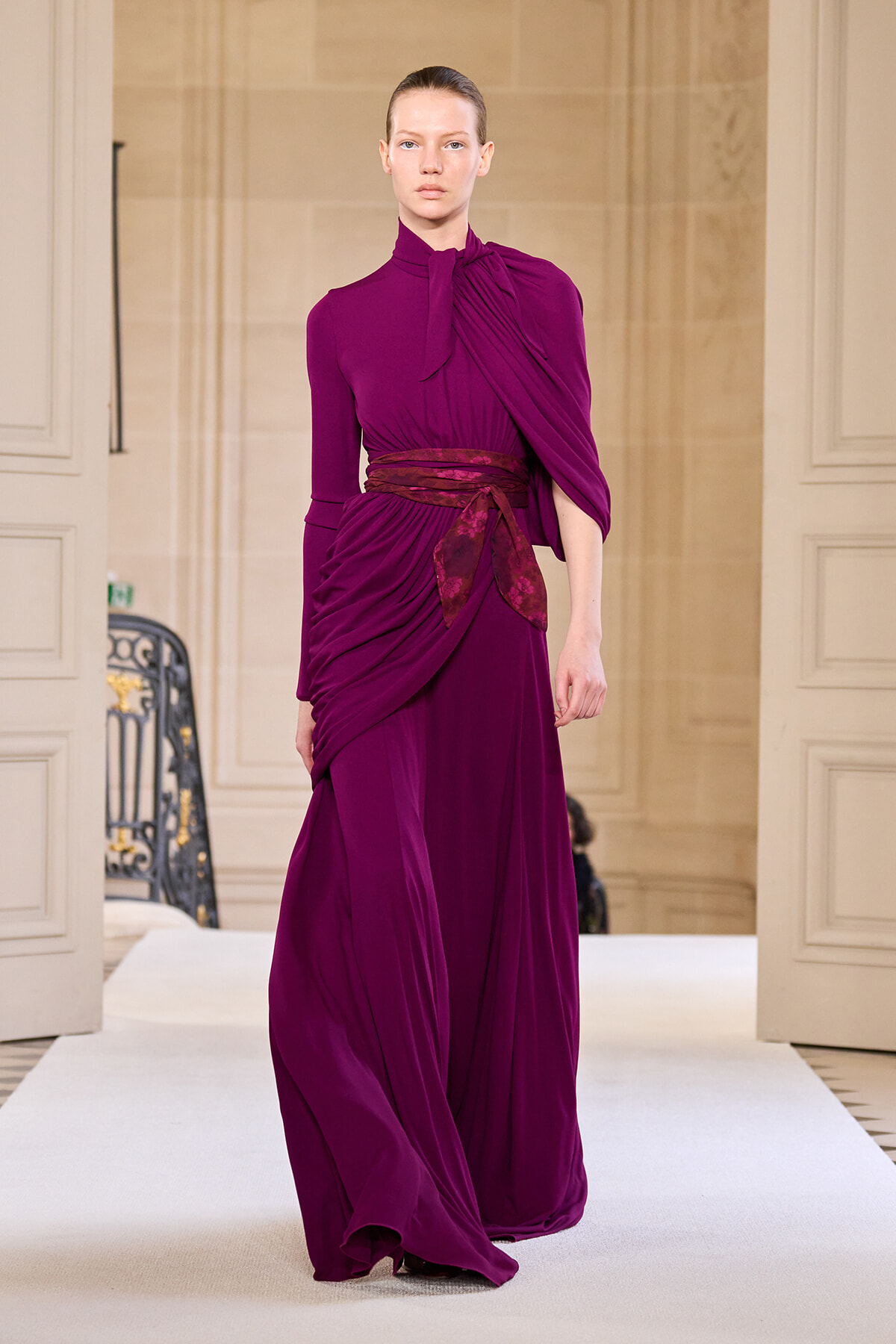 Model walking a runway in a floor-length magenta dress with draped bodice and brown sash tied at the waist.