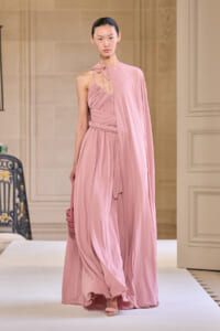 Model walking in a pale pink, one-shoulder halter gown with draped cape and pleated bodice, carrying a small pink handbag on a runway