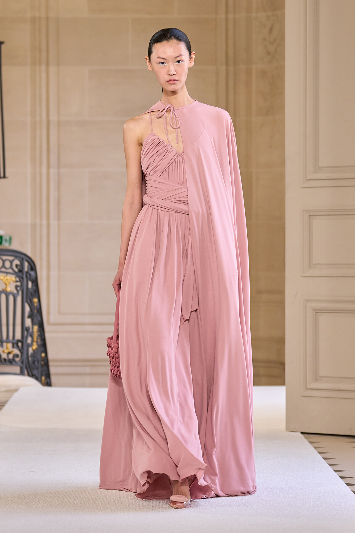 Model walking in a pale pink, one-shoulder halter gown with draped cape and pleated bodice, carrying a small pink handbag on a runway