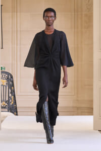 Model walks the runway in a black knee-length dress with a twisted knot at the waist and sheer cape-like sleeves, paired with tall black boots.