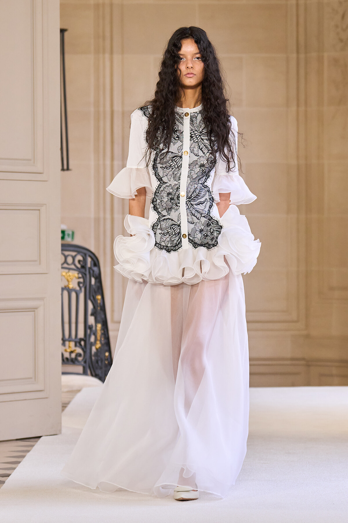 Model walking a runway in a white gown featuring a black lace bodice, sheer puff sleeves, gold buttons, and a voluminous layered skirt in a grand interior hallway.