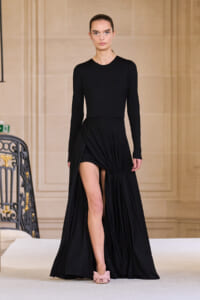 Model walking a fashion runway in a black long-sleeve gown with a high slit, paired with pink bow-front shoes.