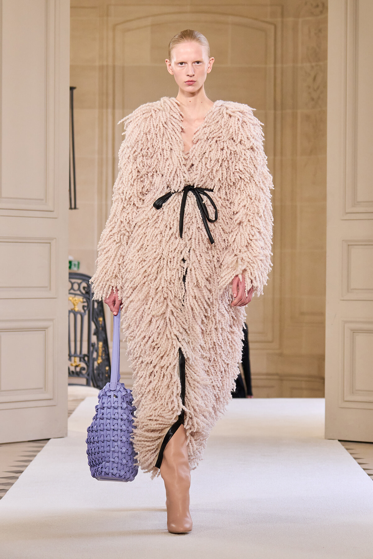 Model walks the runway in a voluminous beige shag coat with a black belt, carrying a lavender woven handbag and wearing nude boots.