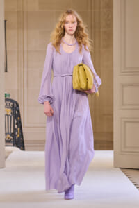 Model on a runway wearing a lavender maxi dress with billowy sleeves and a cinched waist, carrying a mustard-yellow handbag.