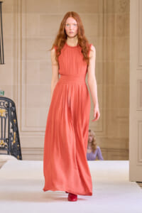 Model walking a runway in a coral pink sleeveless gown with a pleated bodice and cinched waist on a designer catwalk.