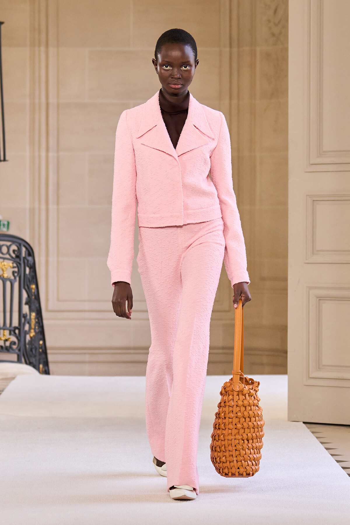 Model walking a runway in a pale pink textured suit with a cropped jacket and wide-leg pants, carrying a woven tan basket tote.