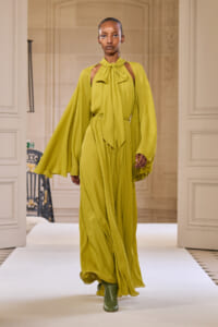 Model walks the runway in a lime-green, floor-length dress with cape sleeves and a neck-tie bow, paired with green boots.