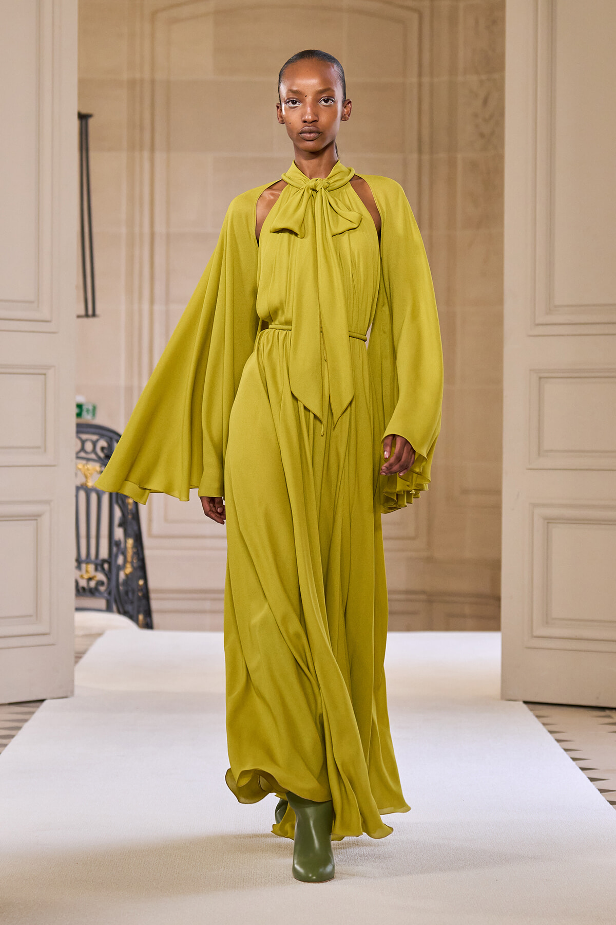 Model walks the runway in a lime-green, floor-length dress with cape sleeves and a neck-tie bow, paired with green boots.