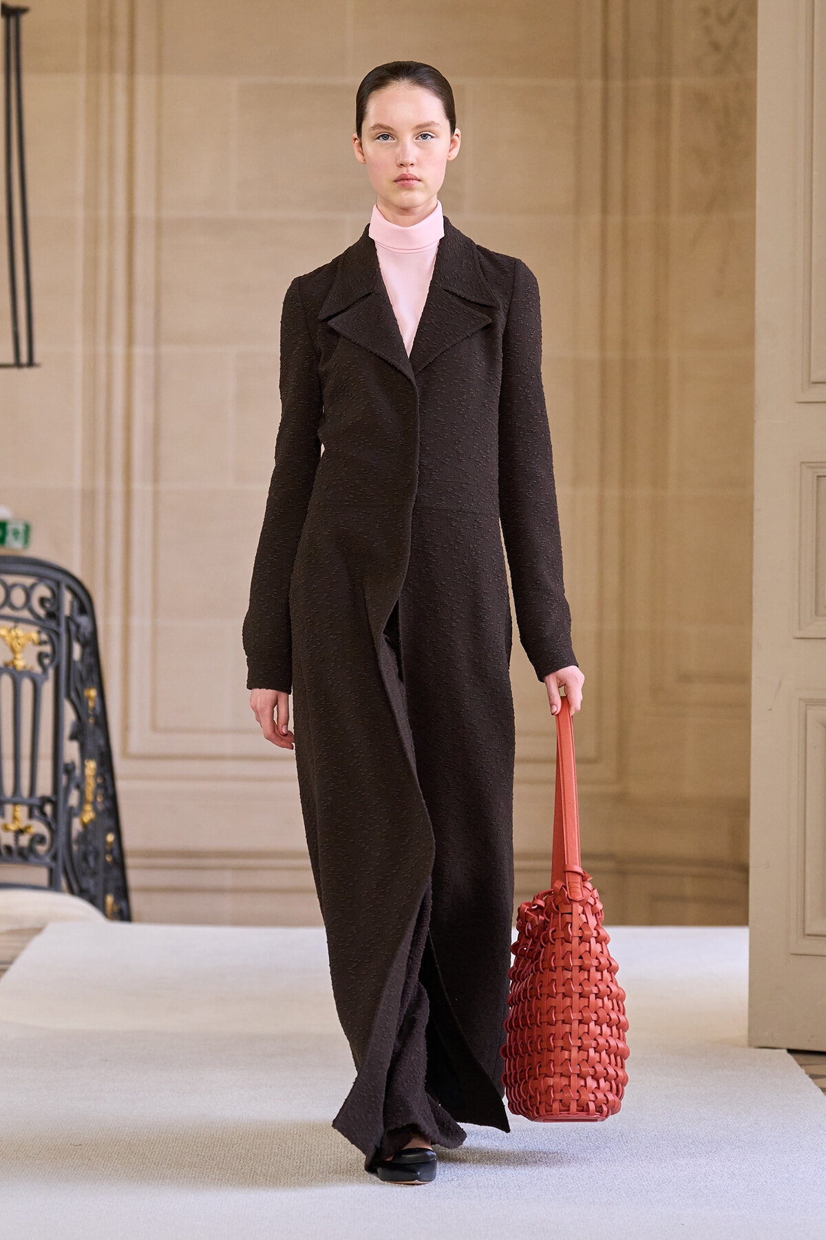 Runway model in a long black textured coat with a pale pink turtleneck, carrying a red woven tote bag.