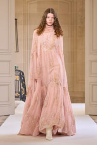 Model walking a runway in a pale pink, floor-length floral gown with sheer sleeves and a high neck, on a white catwalk.