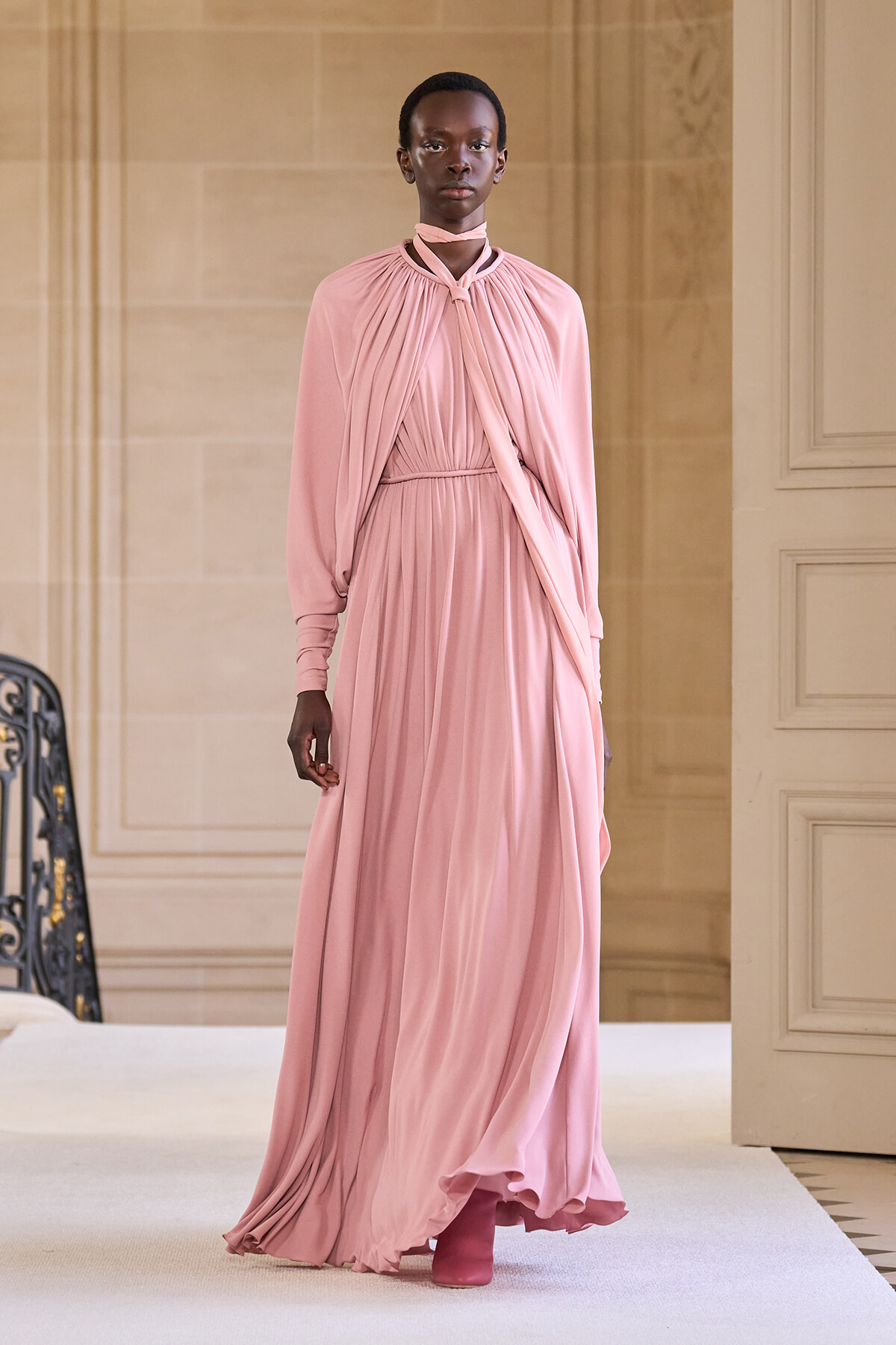 Model walking a runway in a floor-length, soft pink draped gown with cape-like overlay and matching boots in an elegant interior setting