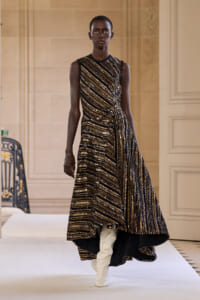 Model walking a runway in a sleeveless, gold-and-black striped sequin gown with a high-low hem and white knee-high boots.