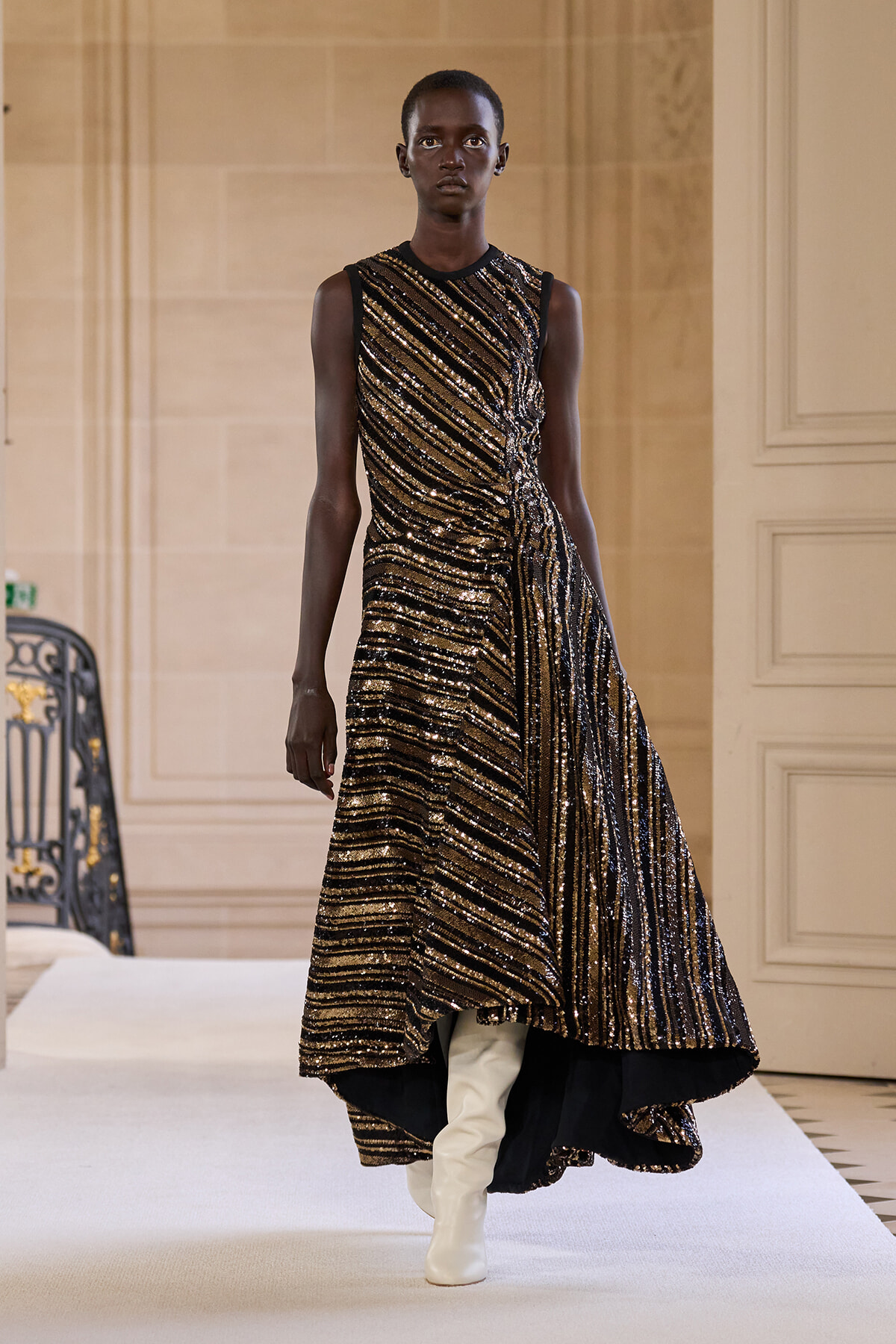 Model walking a runway in a sleeveless, gold-and-black striped sequin gown with a high-low hem and white knee-high boots.