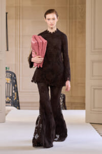 Model walks a runway in a black lace two-piece outfit, holding a pink folded garment under her arm.