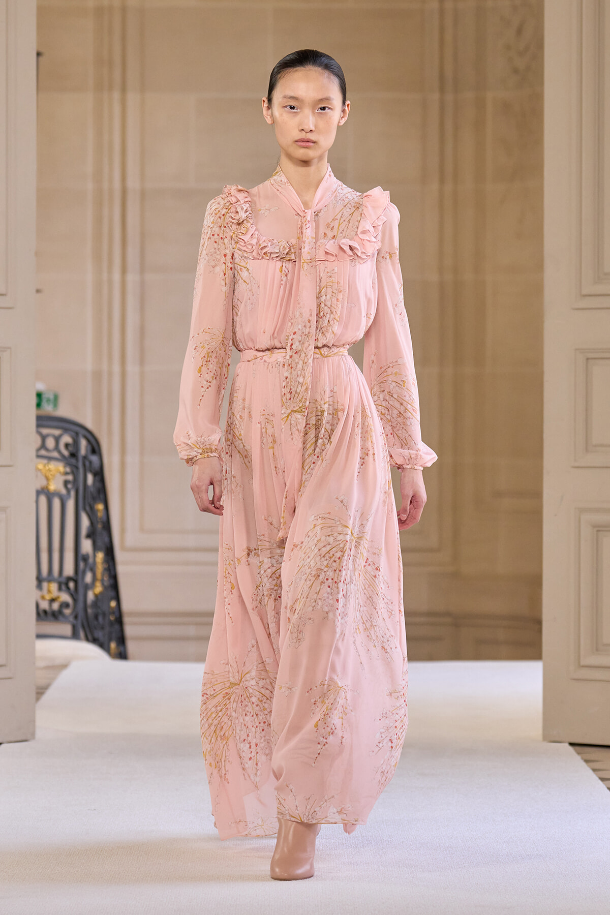 Runway model wearing a pale pink floral maxi dress with ruffled bodice and tied neck.