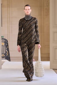 Model on a runway wearing a gold and black diagonal-striped sequined turtleneck top and matching wide-leg pants, carrying a white woven tote.