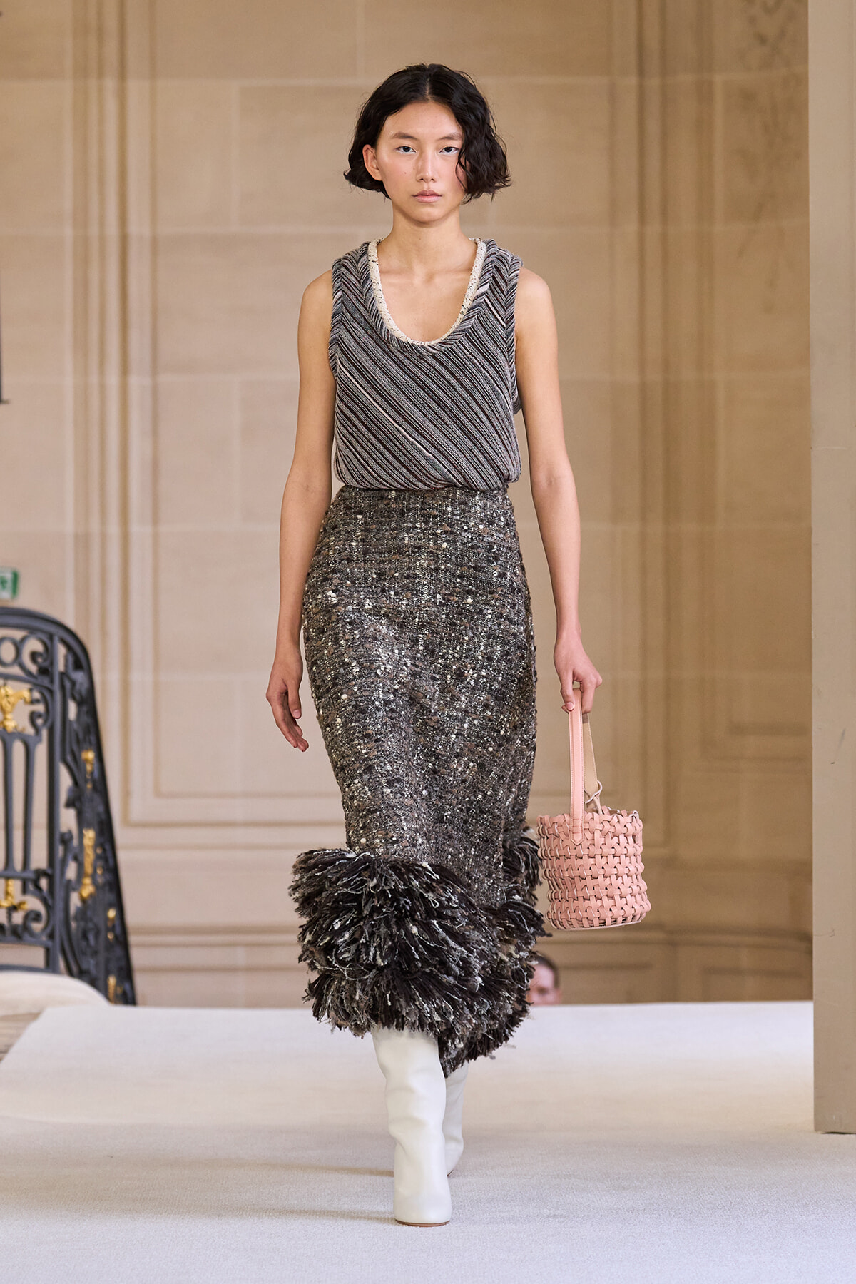 Model walking a runway in a sleeveless striped knit top tucked into a sequined midi skirt with feathered hem, paired with white knee-high boots and a pink woven handbag in a grand interior setting.