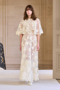 Model on a runway wearing a floor-length ivory lace dress with floral appliqués and scalloped edges.