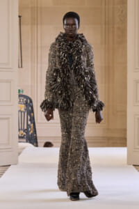 Black model walks a runway wearing a silver-sequined, floor-length gown with a voluminous feathered scarf in an ornate interior setting.
