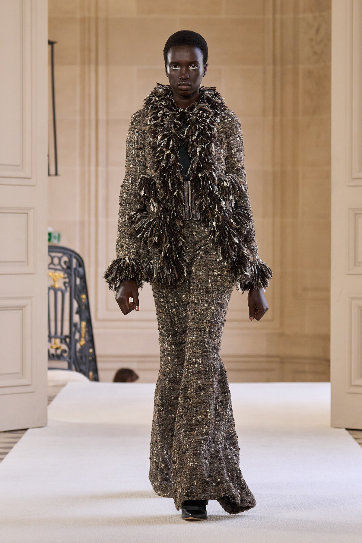 Black model walks a runway wearing a silver-sequined, floor-length gown with a voluminous feathered scarf in an ornate interior setting.
