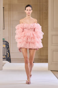 Model walking a runway in a pale pink, off-the-shoulder, multi-layered ruffle dress with matching pink heels.