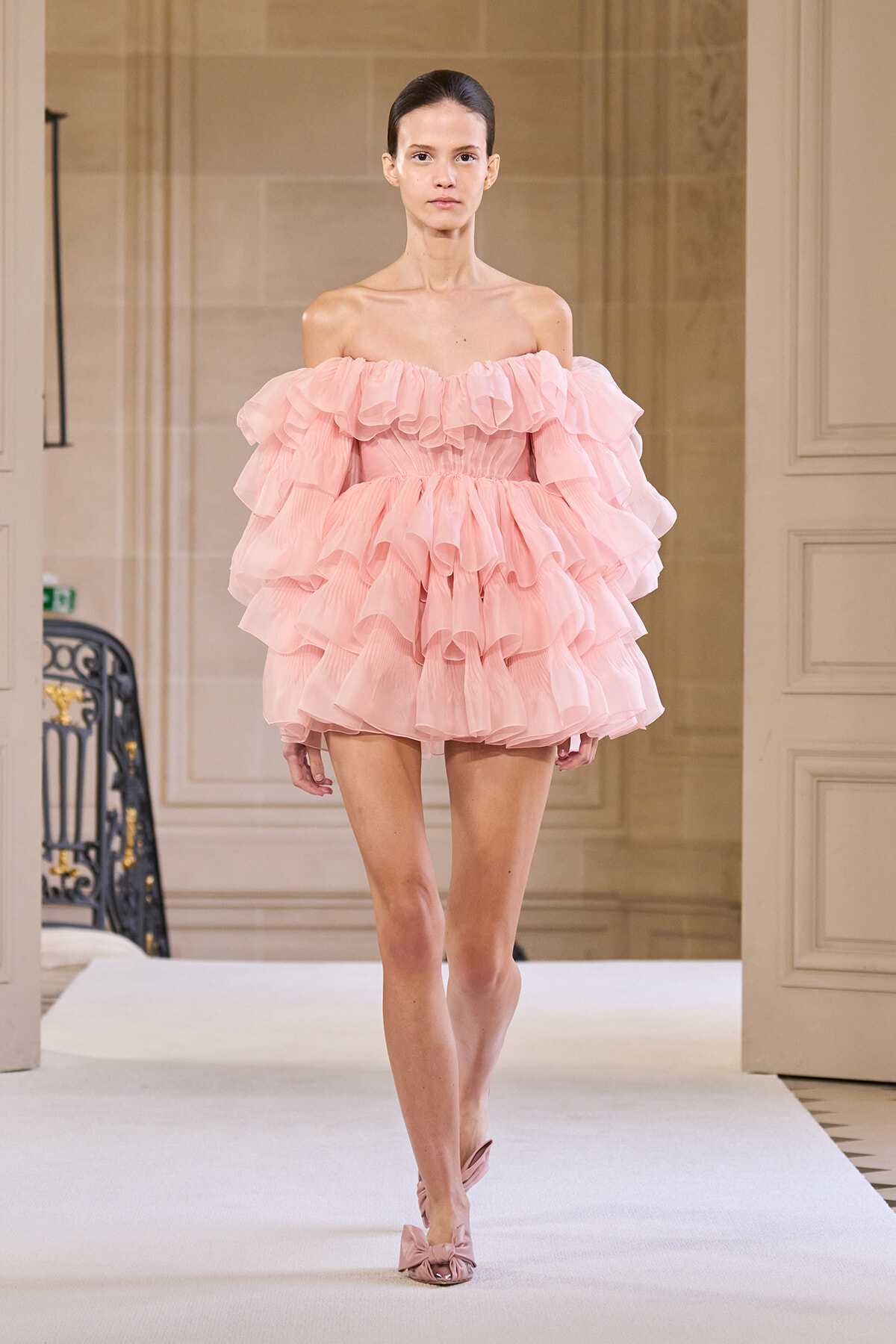 Model walking a runway in a pale pink, off-the-shoulder, multi-layered ruffle dress with matching pink heels.