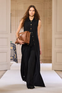 Model walks a runway in a sleeveless black button-front long vest with gold buttons, carrying a brown leather handbag under an ornate interior backdrop.