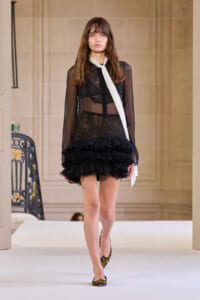 Model walking a runway in a sheer black lace dress with long sleeves, tiered ruffled mini-skirt, and a white neck scarf; pointed floral-patterned heels visible.
