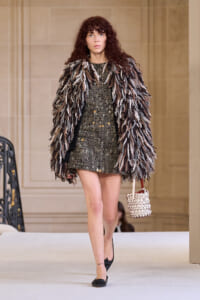 Model on a runway wearing a chunky shaggy fur coat over a metallic mini dress, carrying a white woven handbag.