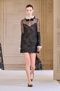 Model on a runway wearing a black lace mini dress with sheer sleeves and a white ruffled collar.