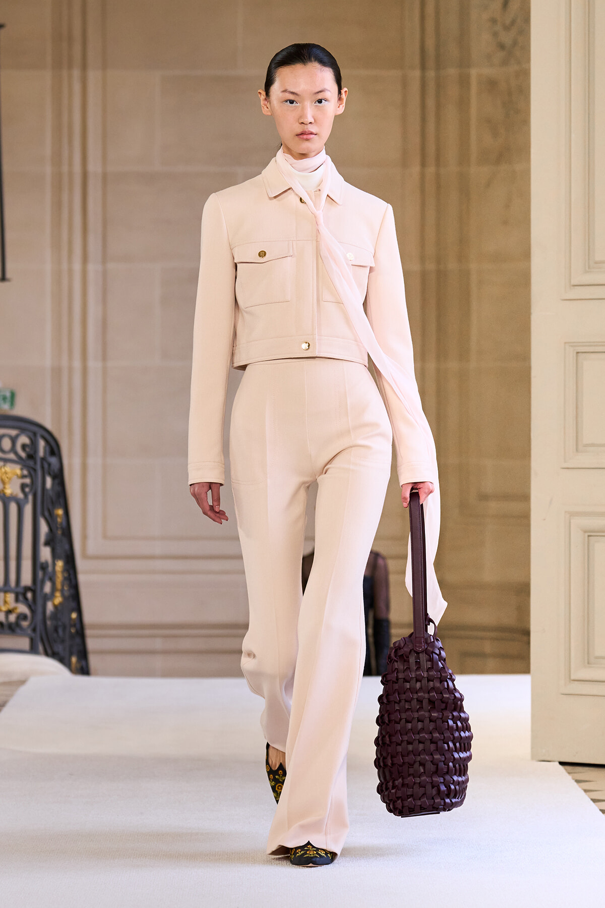 Model walking the runway in a pale beige suit with scarf, carrying a dark purple woven tote bag.