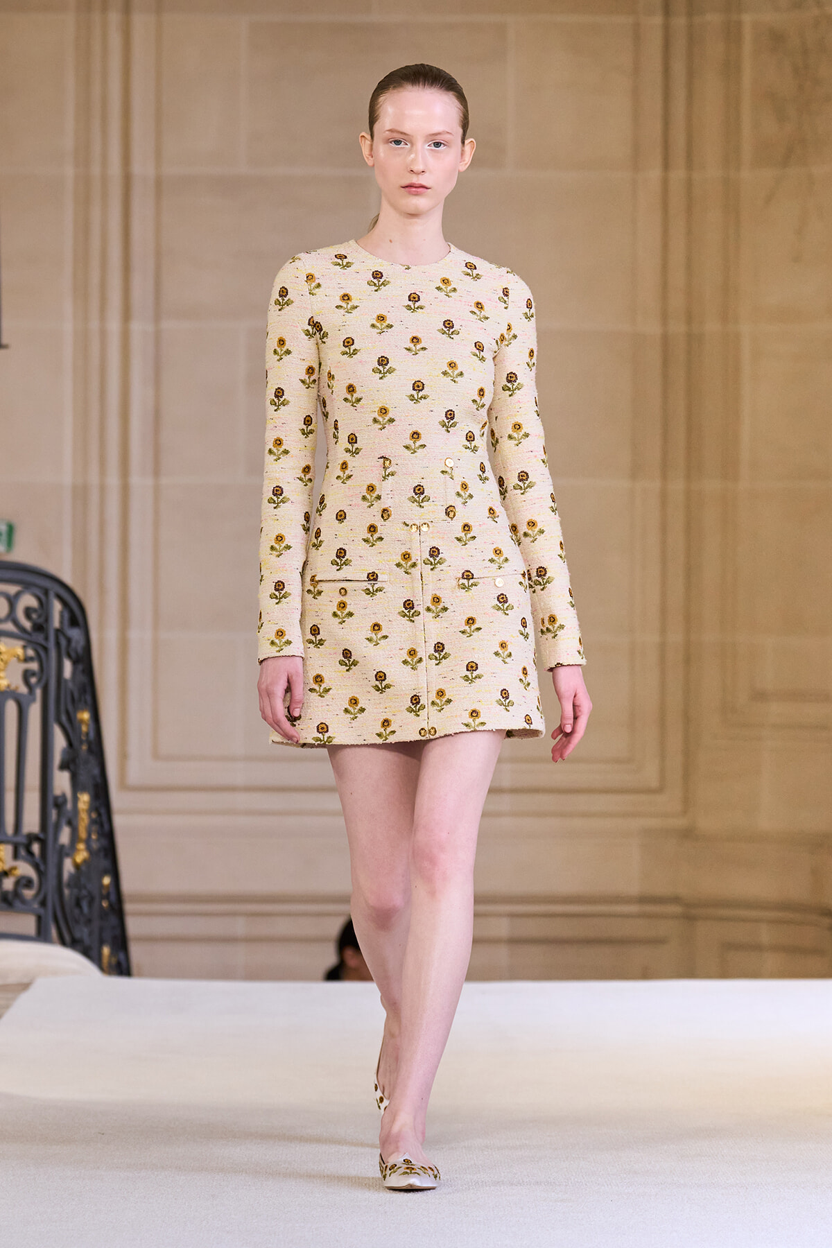 Model walking a beige floral knit mini dress down a runway, matching flats with the same pattern.