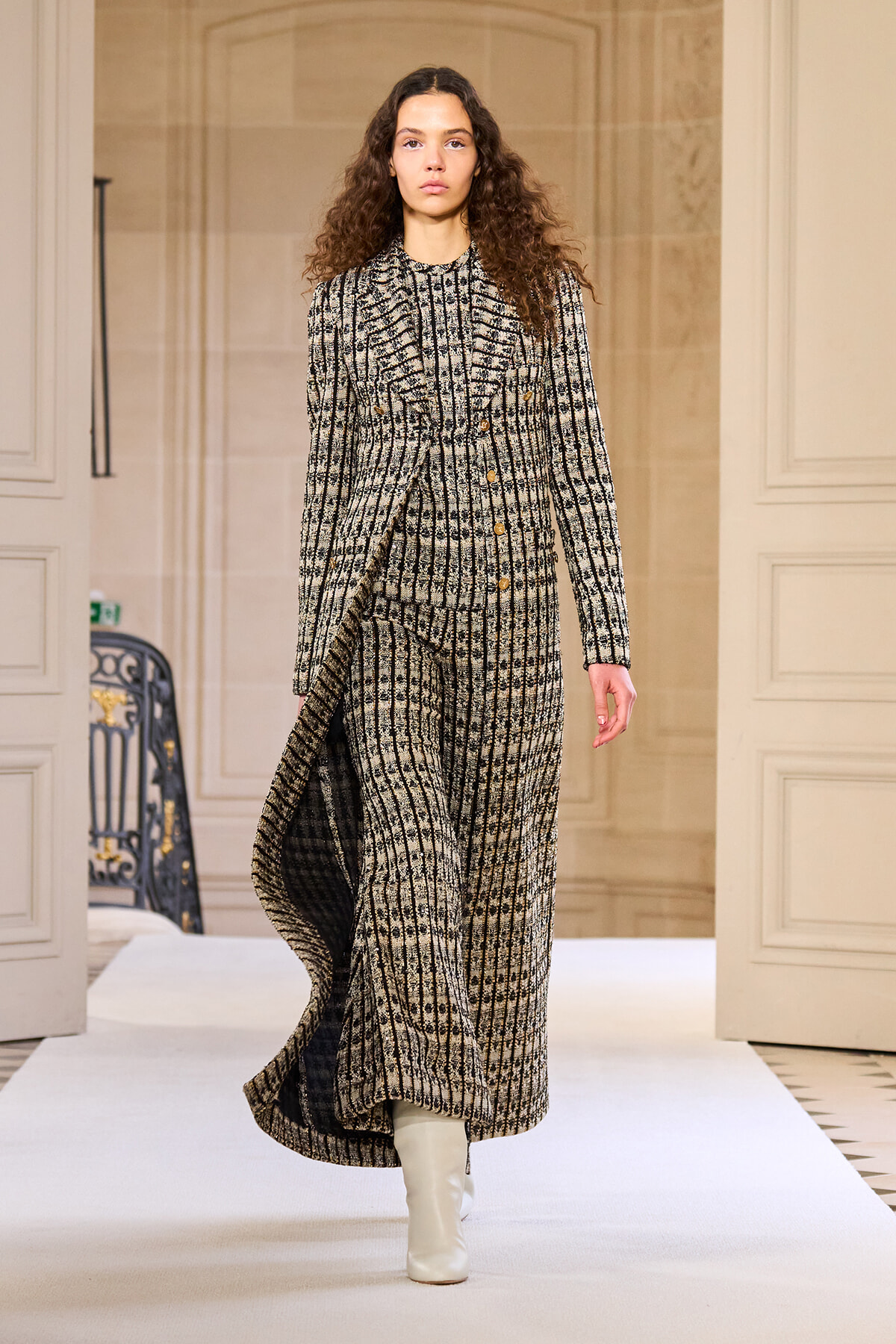 Model walks a runway in a long black-and-white plaid tweed coat with gold buttons, paired with white knee-high boots in a beige set.