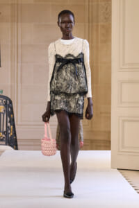 Model walking a runway in a cream knit top with a black lace overlay and tied bow, carrying a pink woven handbag.
