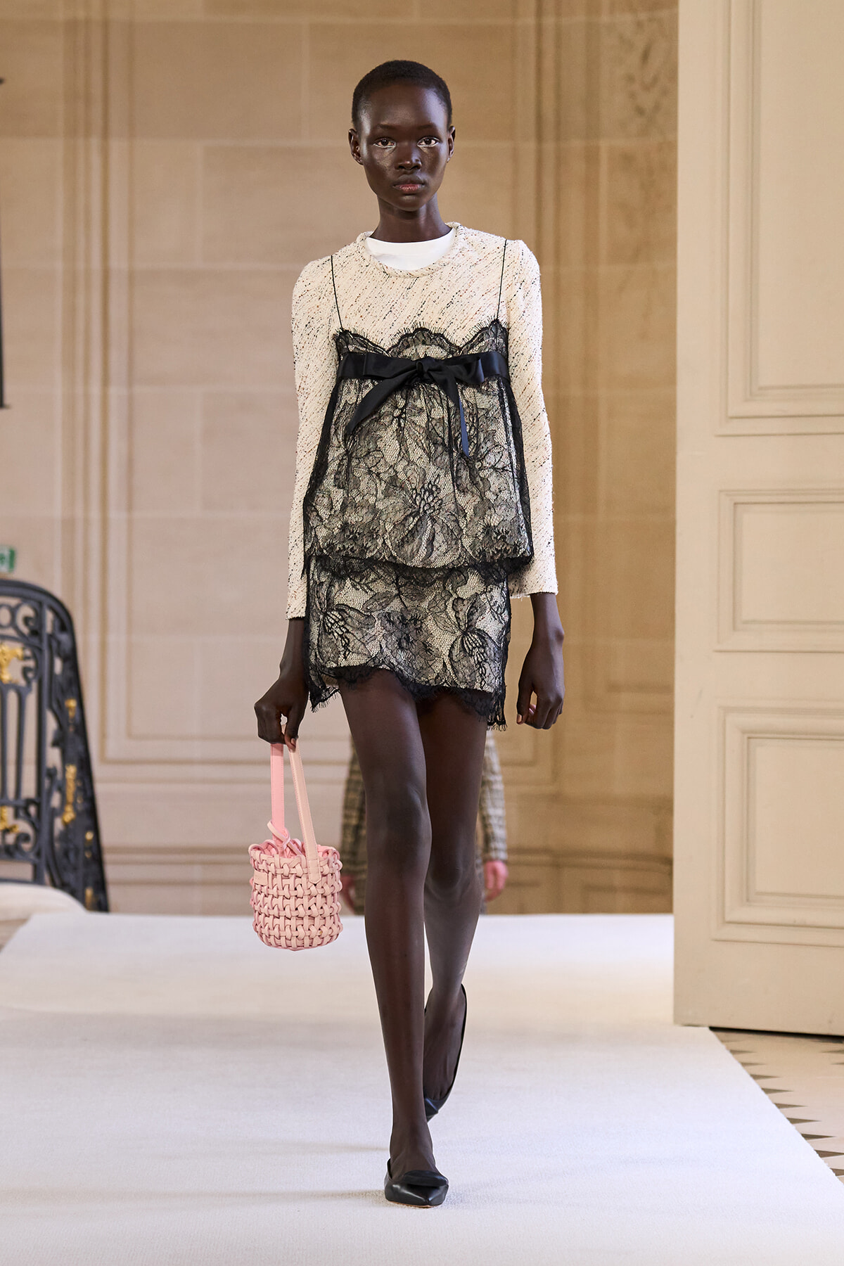 Model walking a runway in a cream knit top with a black lace overlay and tied bow, carrying a pink woven handbag.