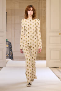 Model walks a runway in a cream floral print knit jumpsuit on a white catwalk stage.