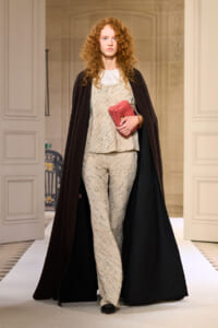 Model walking runway wearing beige speckled pantsuit with a long dark brown coat, holding a pink clutch.